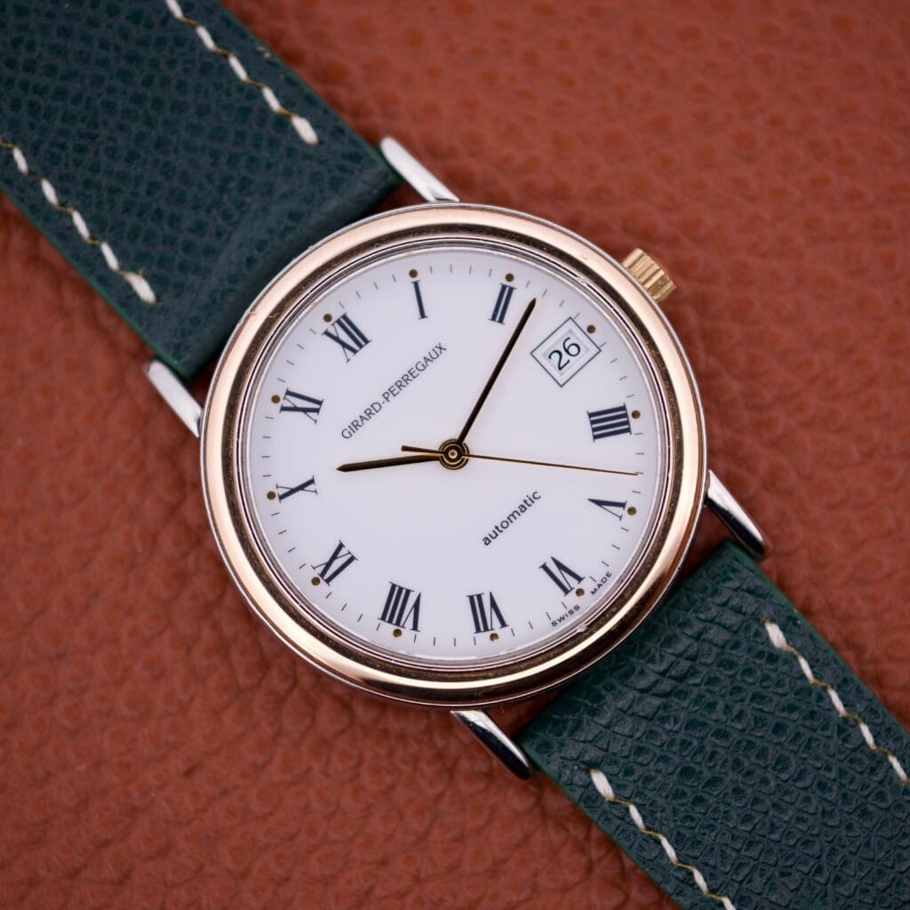 The Girard Perregaux Automatic 4797 is a round wristwatch with a white dial, Roman numerals, automatic movement, date display, and a green leather strap on brown leather.