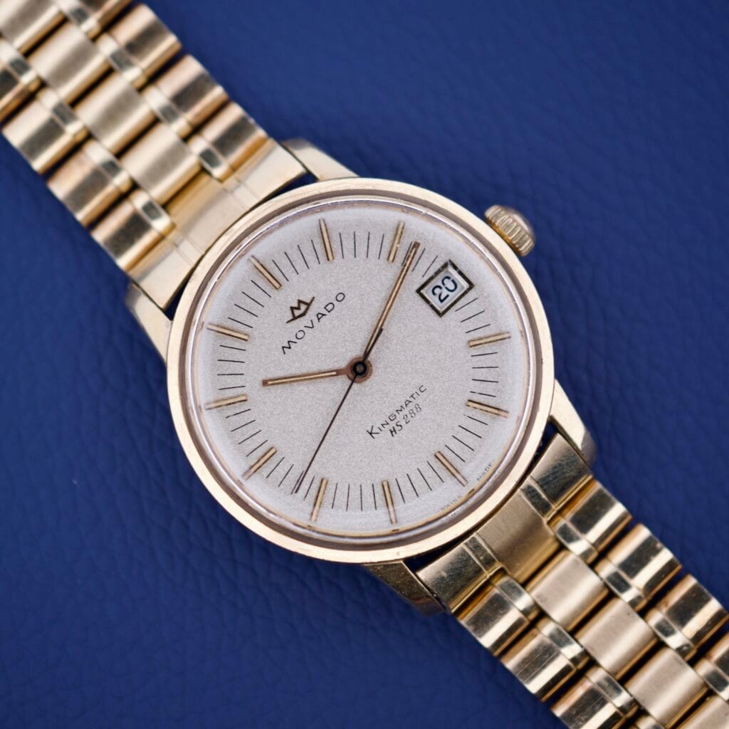 Movado Kingmatic HS 288 Automatic wristwatch with date display on a blue textured surface.