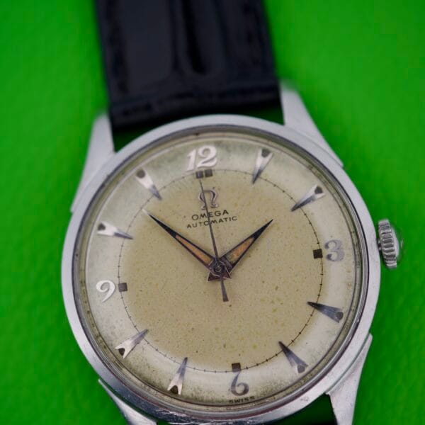An Omega Automatic Bumper 2635 Original Strap wristwatch with a yellowed dial and original black leather strap, showcased against a bright green background.