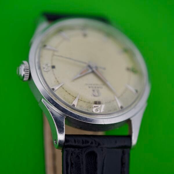 Close-up of an Omega Automatic Bumper 2635 Original Strap wristwatch featuring a cream dial and original black leather strap, displayed against a green background.