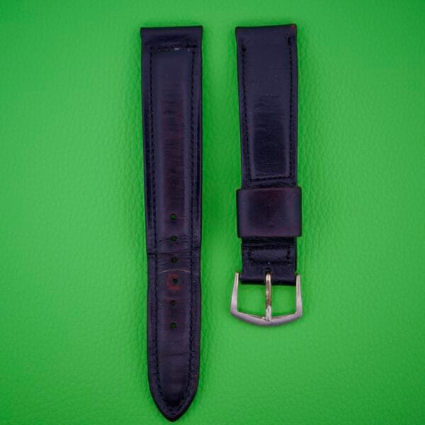 Two pieces of the Omega Automatic Bumper 2635 Original Strap, made from black leather with a buckle, are displayed on a bright green textured background.