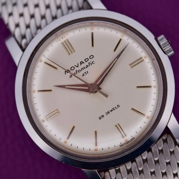 Close-up of a Movado Automatic 431 Stainless Steel wristwatch with a mesh band, displayed on a purple background.