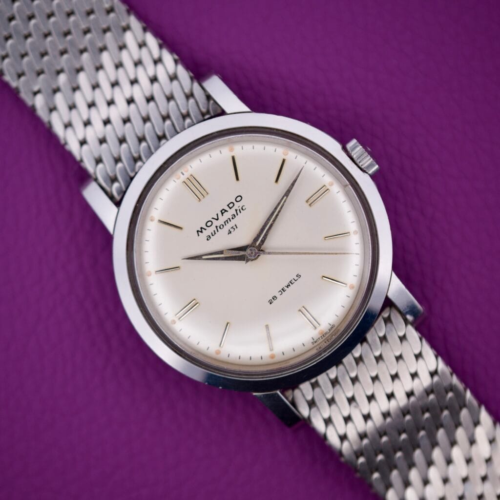 The Movado Automatic 431 Stainless Steel wristwatch with a mesh band is displayed on a textured purple background.