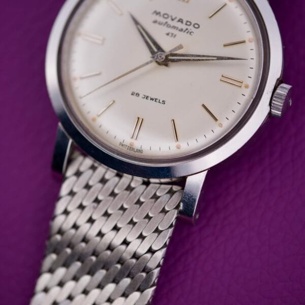 Close-up of the Movado Automatic 431 Stainless Steel wristwatch with mesh band, shown on a purple textured surface.
