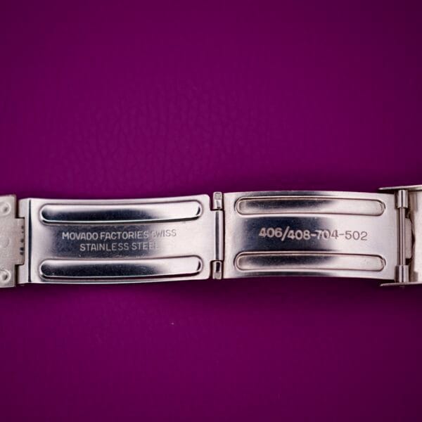 Silver stainless steel watch clasp marked MOVADO FACTORIES SWISS, shown on a purple background—an elegant accessory for your Movado Automatic 431 Stainless Steel.