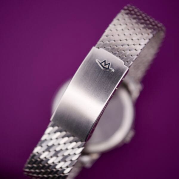 Close-up of the Movado Automatic 431 Stainless Steel watch band with a crown logo clasp, displayed against a purple background.