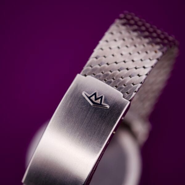 Close-up of the Movado Automatic 431 Stainless Steel watchband clasp featuring a crown logo, set against a blurred purple background.