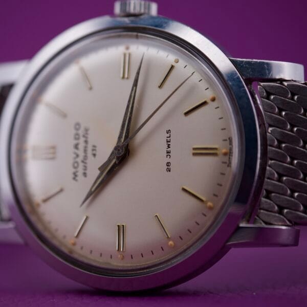 Close-up of a Movado Automatic 431 Stainless Steel wristwatch with a white face and silver band on a purple background.