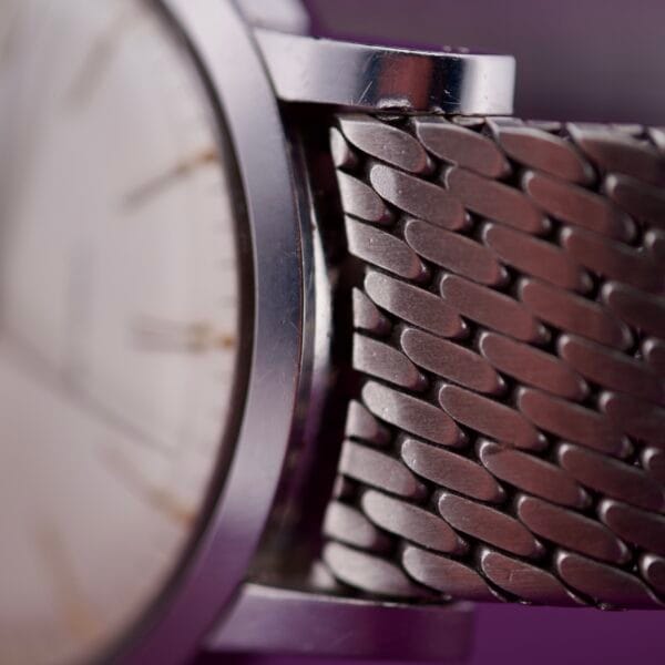 Close-up of the Movado Automatic 431 Stainless Steel watch with a metal mesh band, set against a blurred purple background.