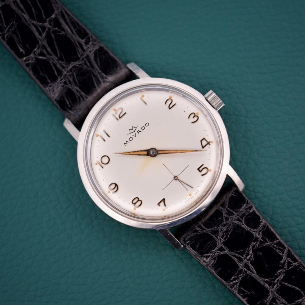 A Movado Sub Sea Arabic Numerals wristwatch with a black leather strap rests on a textured green background.