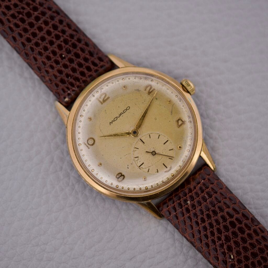 Movado Jumbo Manual Wind Sub Seconds watch in gold with a brown leather strap, featuring a sub seconds dial, displayed on a light textured background.