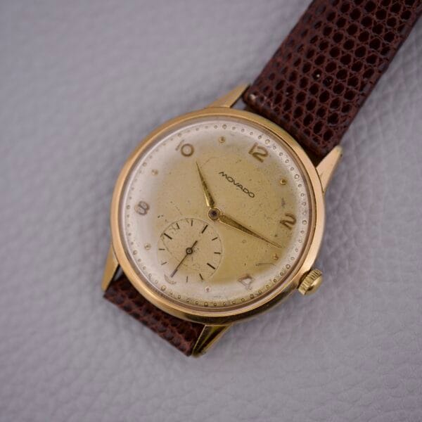 A Movado Jumbo Manual Wind Sub Seconds wristwatch with a gold case and brown leather strap rests on a gray textured surface.