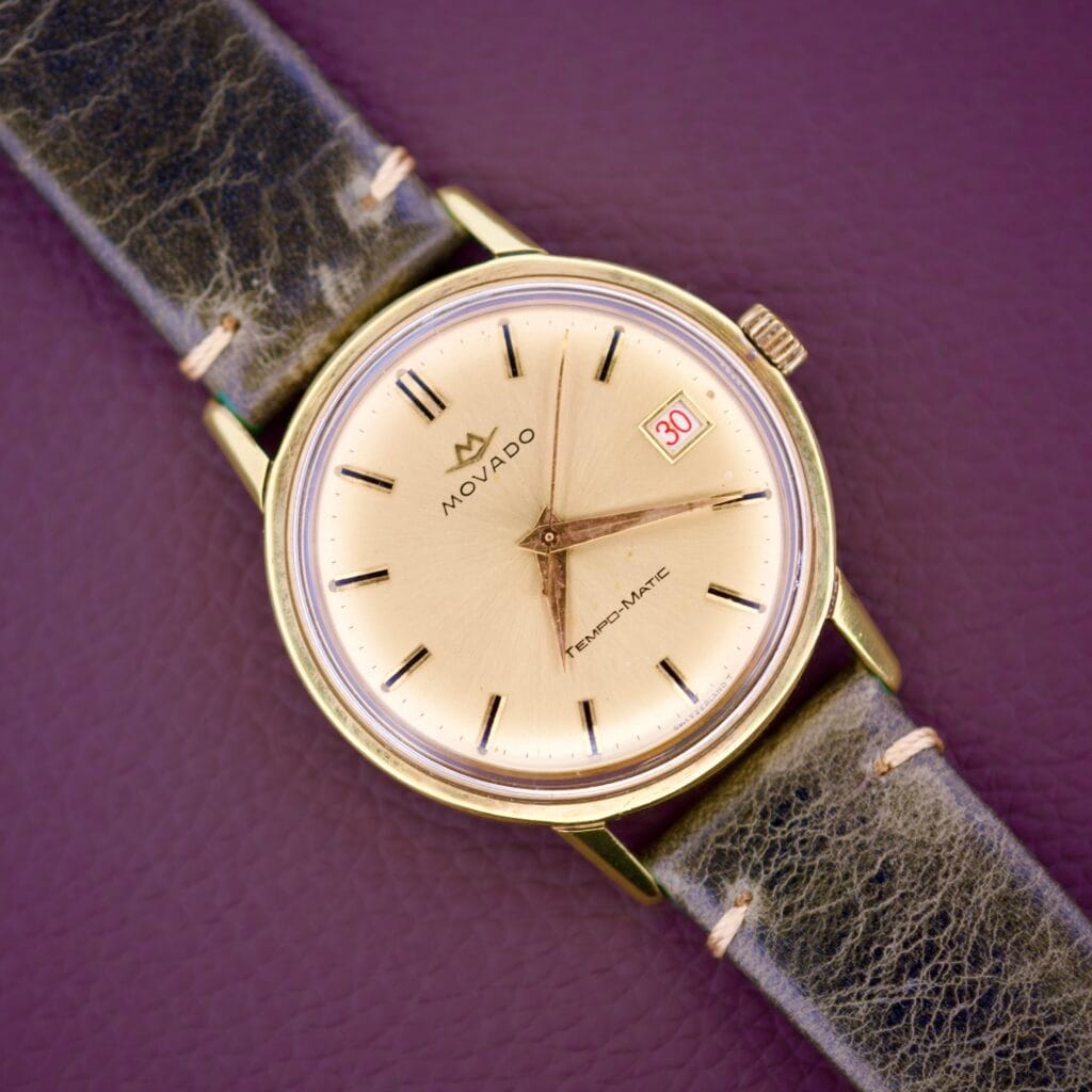 The Movado Tempo-Matic Sub Sea Roulette Date is a gold wristwatch with a black leather strap and a Roulette Date display showing 30 on a purple background.