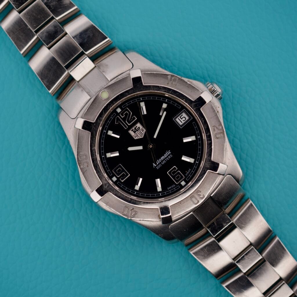 The Tag Heuer 2000 Automatic Diver WN2111 wristwatch with a black dial rests on a turquoise textured surface, highlighting its classic design.