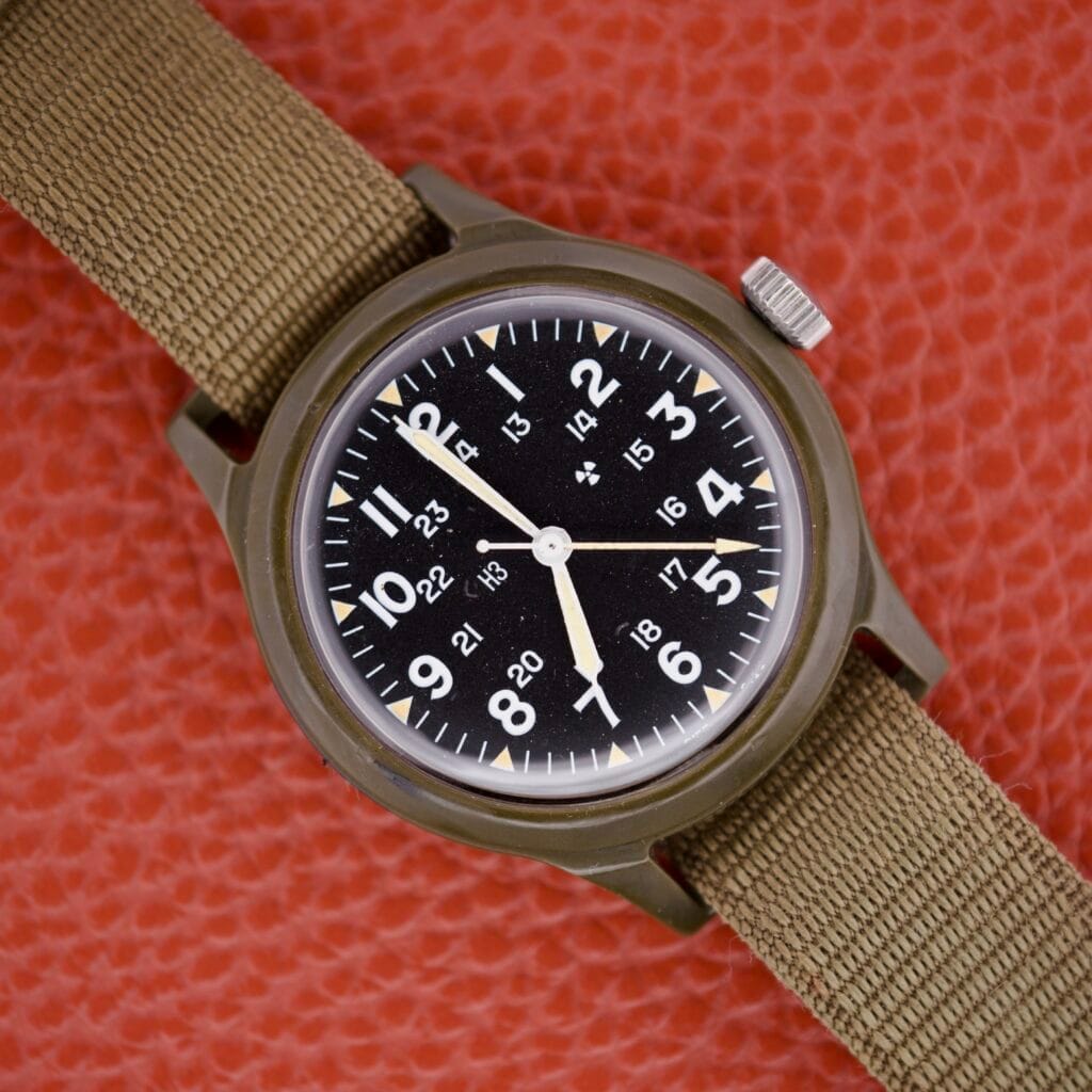 The Benrus Mil-W-46374A US Military 1975 analog watch with a khaki strap and bold black dial lies on a textured orange surface, showcasing military-inspired design.