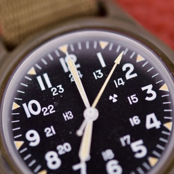 Close-up of a Benrus Mil-W-46374A US Military 1975 black analog watch face, with white numbers and tan hour and minute hands, echoing the classic design of vintage military timepieces.