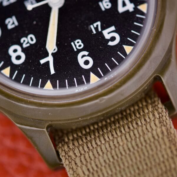 Close-up of the Benrus Mil-W-46374A US Military 1975 wristwatch with a khaki fabric strap and black dial, featuring numbers 6 to 8—an iconic design based on the original 1975 military specifications.