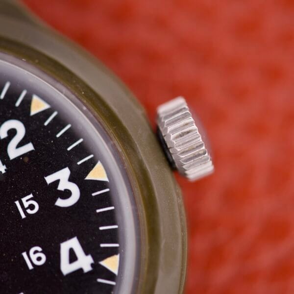 Close-up of the Benrus Mil-W-46374A US Military 1975 watch face, featuring numbers 2 to 4 and the crown, set against a textured red background.