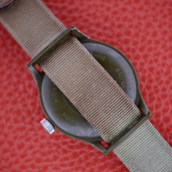 Back of a green Benrus Mil-W-46374A US Military 1975 wristwatch with tan nylon strap on a textured red surface.