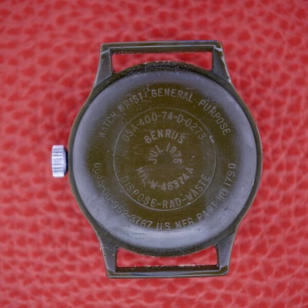 Back of a Benrus Mil-W-46374A US Military 1975 wristwatch on a red textured surface, displaying engraved serial info and Mil-W-46374A markings.