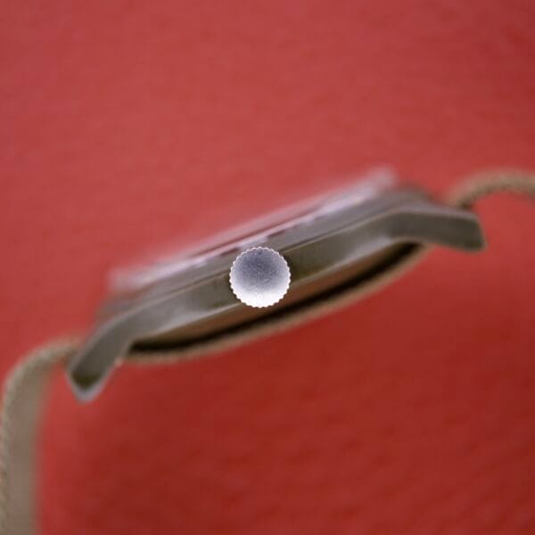 Close-up of the Benrus Mil-W-46374A US Military 1975 watch crown, set against a blurred red background with parts of the strap visible on both sides.