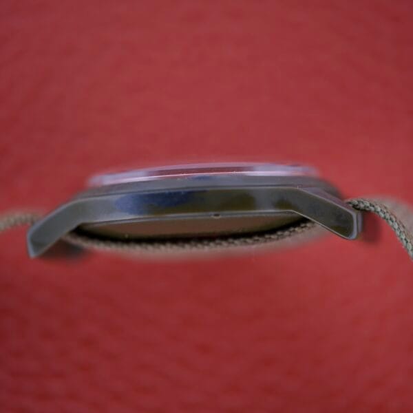 Close-up side view of the Benrus Mil-W-46374A US Military 1975 wristwatch with a fabric strap, set against a textured red background.