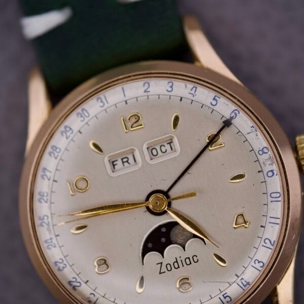 The Zodiac Triple Calendar Moonphase Cal. 12S wristwatch showcases a gold case, triple calendar with moonphase, day, and date displays, and is paired with a green leather strap.