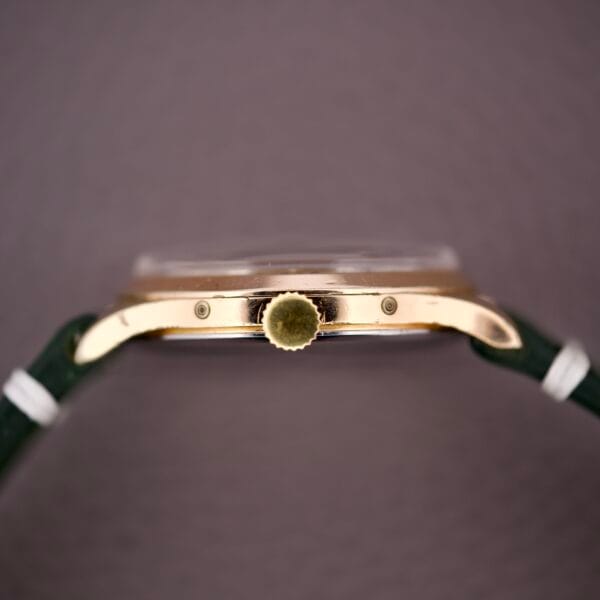 Side view of the Zodiac Triple Calendar Moonphase Cal. 12S, a gold wristwatch with a green strap, textured crown, and elegant triple calendar complication, set against a blurred background.