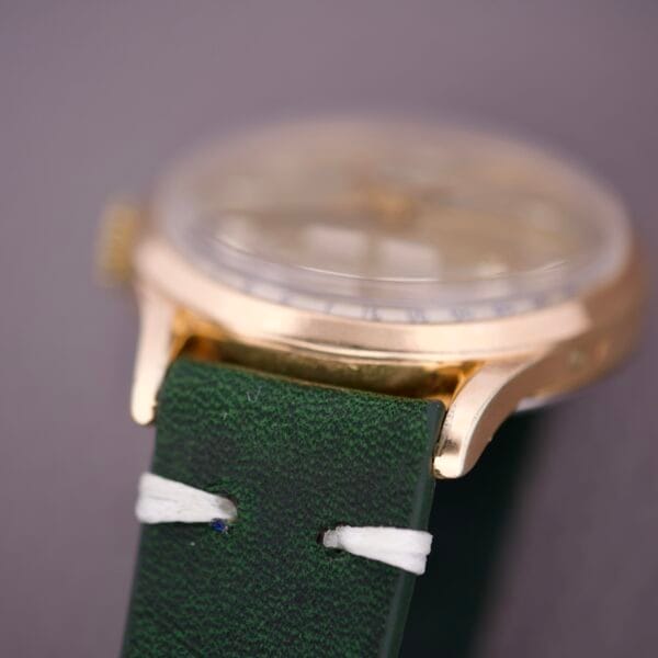 Close-up of the Zodiac Triple Calendar Moonphase Cal. 12S gold wristwatch, showcasing a green leather strap with white stitching and a triple calendar display, set against a blurred background.