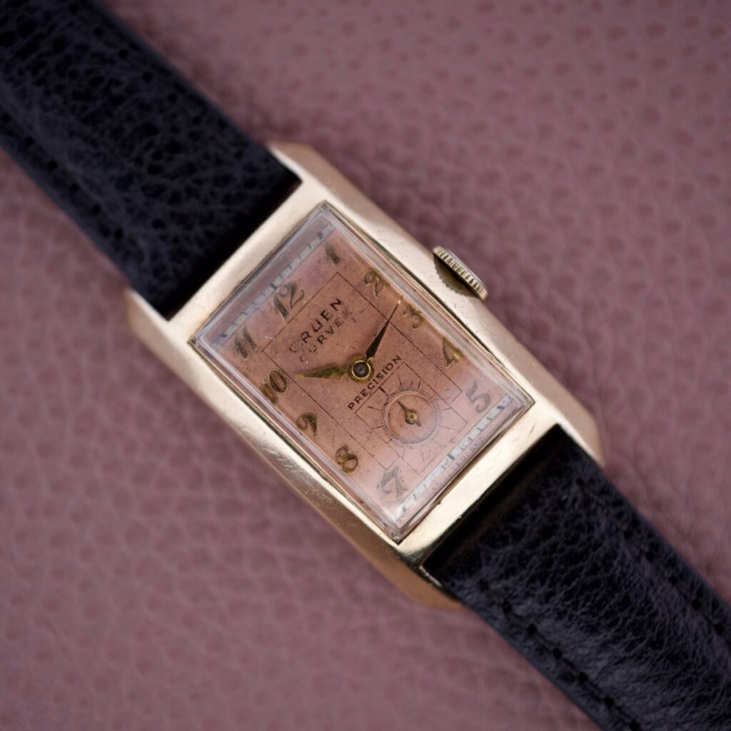 A vintage Gruen Curvex Precision Salmon Dial wristwatch with expert craftsmanship, a rectangular gold case, and black leather strap displayed on a pink surface.