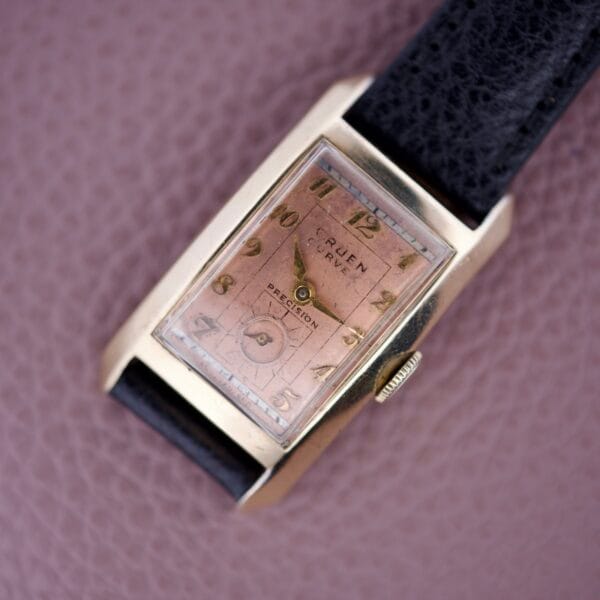 The Gruen Curvex Precision Salmon Dial wristwatch, featuring a rectangular case and black leather strap, is displayed on a textured mauve background.