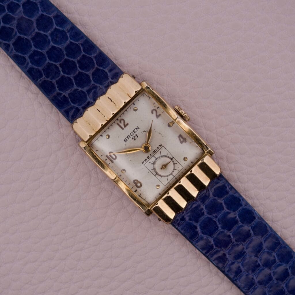 The Gruen 21 Precision Tank USA Cal. 335 is a rectangular vintage wristwatch with a blue textured leather strap, shown on a light textured surface.