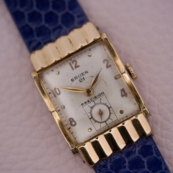 Gruen 21 Precision Tank USA Cal. 335 wristwatch with a blue strap and rectangular Art Deco face, shown on a light textured surface.