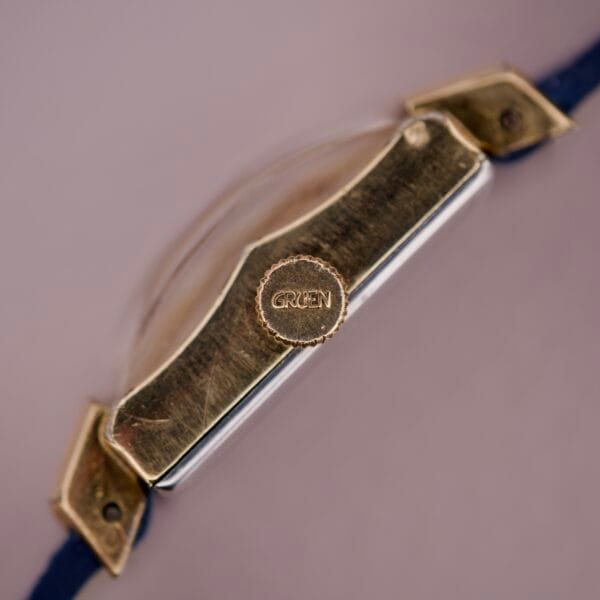 Close-up side view of a Gruen 21 Precision Tank USA Cal. 335 watch in gold with a blue strap, engraved crown, and Gruen 21 Cal. 335 movement.