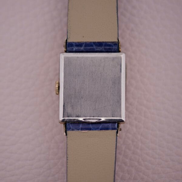 A Gruen 21 Precision Tank USA Cal. 335 wristwatch with a blank square metal face and blue leather strap rests on a textured surface, epitomizing vintage elegance.