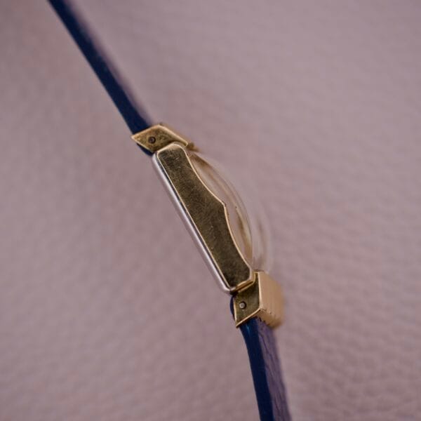 Close-up of a Gruen 21 Precision Tank USA Cal. 335 gold wristwatch case with a blue strap, set on a textured light pink surface.