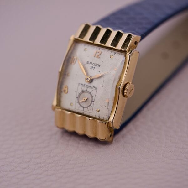 Gruen 21 Precision Tank USA Cal. 335 gold vintage wristwatch with a blue leather strap displayed on a light textured surface.
