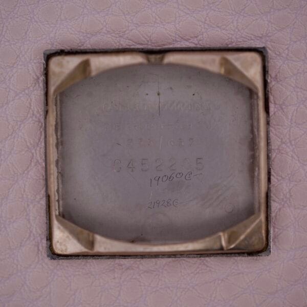 Close-up of a metallic tag engraved with numbers and text referring to Gruen 21 Precision Tank USA Cal. 335, mounted on a textured, light pink surface.