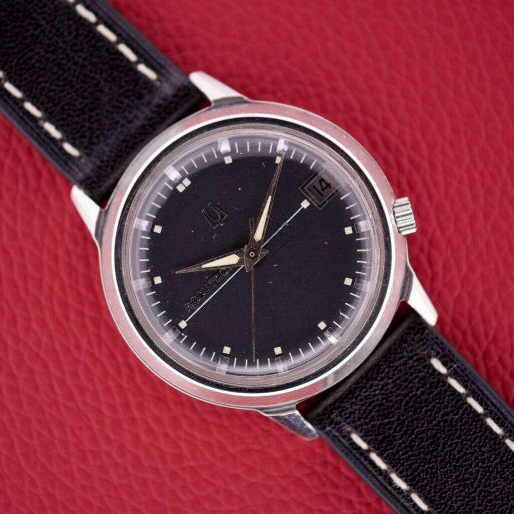 The Bulova Accutron 218 Black Dial wristwatch features a black dial and strap, silver casing, and date display, showcased on a red textured background.