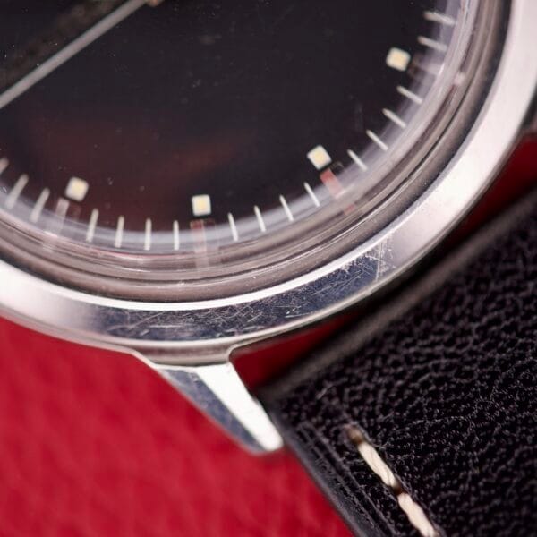 Close-up of a Bulova Accutron 218 Black Dial wristwatch with a silver case and black leather strap on a red textured surface.