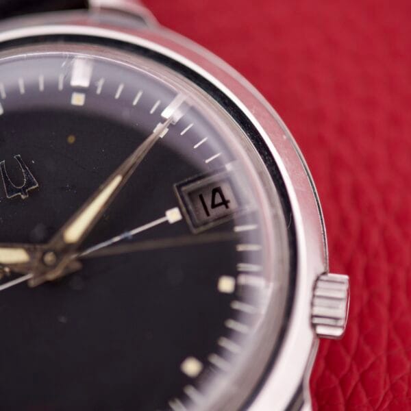 Close-up of the Bulova Accutron 218 Black Dial wristwatch, displaying the date “14” against a textured red background.