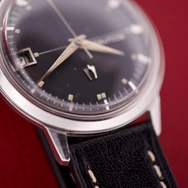 Close-up of the Bulova Accutron 218 Black Dial wristwatch with a leather strap, displayed on a red background.