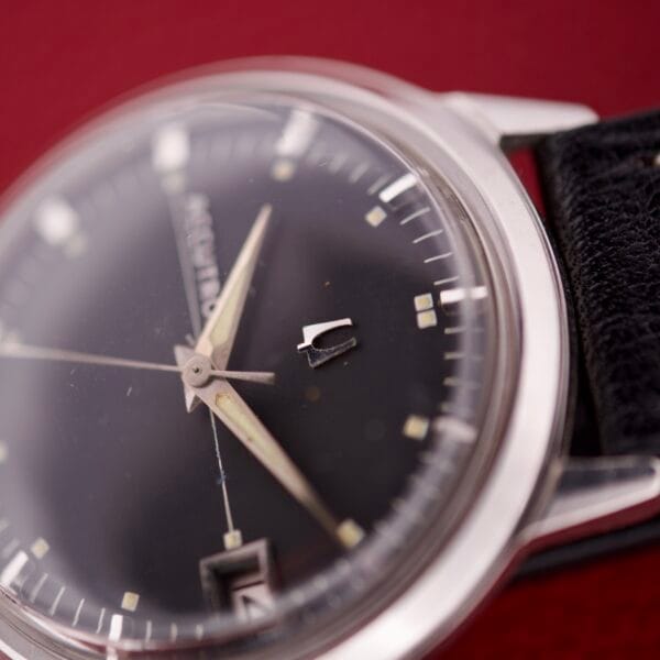 Close-up of the Bulova Accutron 218 Black Dial wristwatch, featuring a sleek black dial and silver case against a vibrant red background.