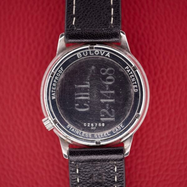 Back of a Bulova Accutron 218 Black Dial watch engraved with CHL 12-14-68, shown on a red textured background to emphasize its classic design.