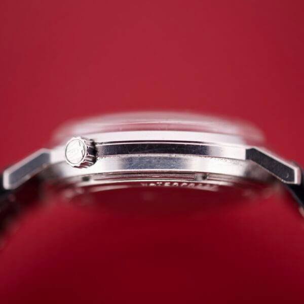 Close-up side view of the Bulova Accutron 218 Black Dial wristwatch with a silver case set against a red background.