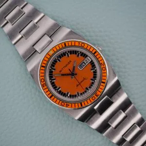 A Wittnauer Diver Bakelite Bezel 1002-W100 with an orange dial, silver strap, and day-date display rests on a textured surface, highlighting its bold design and classic appeal.