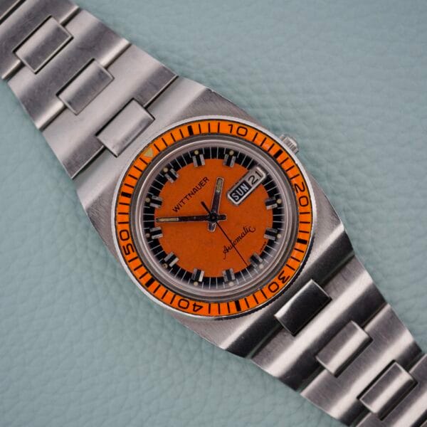 A Wittnauer Diver Bakelite Bezel 1002-W100 with an orange dial, silver strap, and day-date display rests on a textured surface, highlighting its bold design and classic appeal.