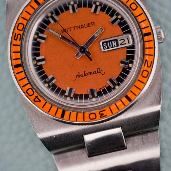 Close-up of a Wittnauer Diver Bakelite Bezel 1002-W100 wristwatch with an orange dial, stainless steel band, and day-date display.