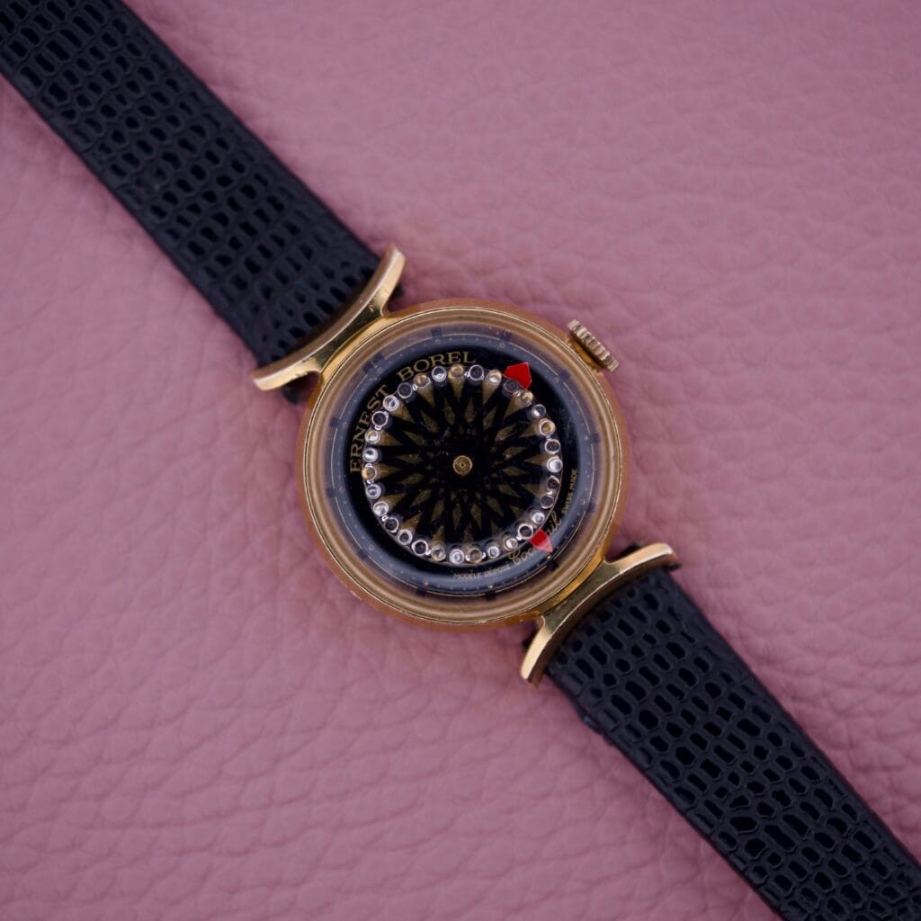An Ernest Borel Kaleidoscope Cocktail 17 Jewels gold wristwatch with a black leather strap is displayed elegantly on a pink textured surface.