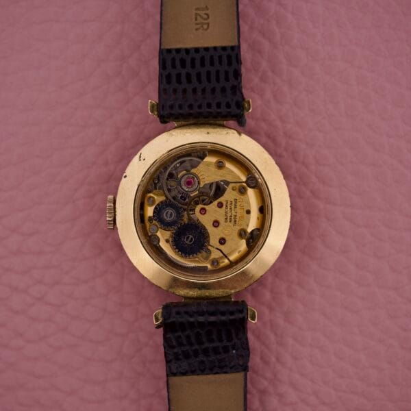 Back view of the Ernest Borel Kaleidoscope Cocktail 17 Jewels gold wristwatch, showing its mechanical gears, displayed on a pink textured surface.